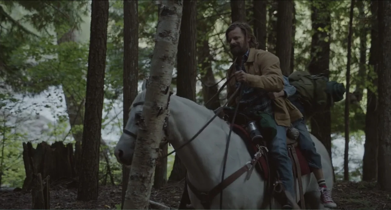 Cowboys (2014) movie still: medium — A dense forest path with tall trees and thick vegetation surrounds the horse riders. A bearded man sits atop a…