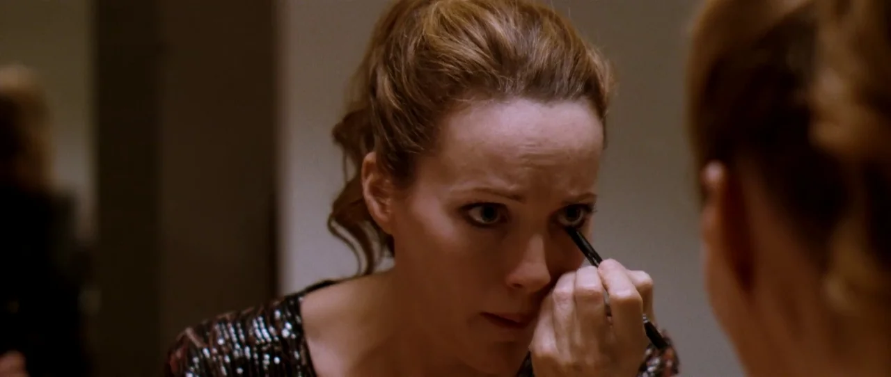 Little Birds (2011) movie still: closeup — A woman stands before a mirror in a dimly lit bathroom setting. She applies black eyeliner to her lower…