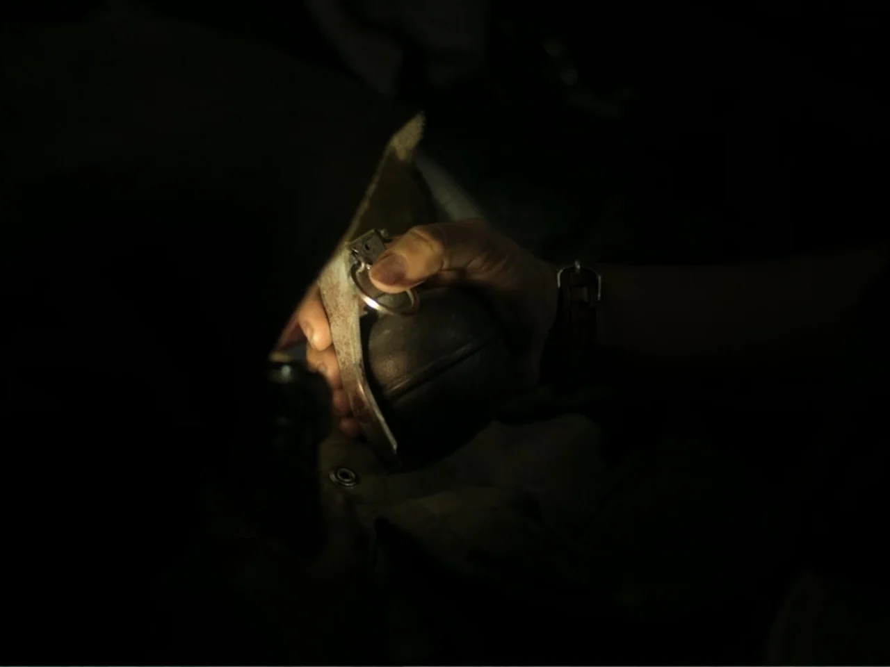November (2025) movie still: closeup — A dimly lit interior space contains a dark metallic object. A hand holds a metal grenade pin in focus.