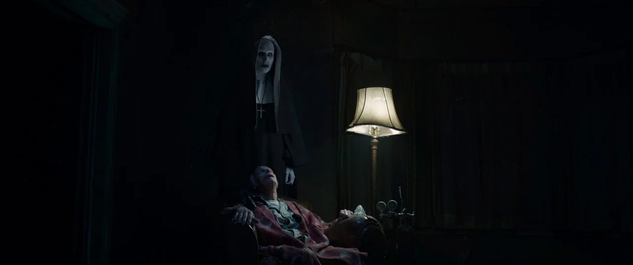The Conjuring 2 (2016) movie still: medium — A dimly lit room with heavy curtains and a glowing floor lamp. A nun stands behind an elderly man…
