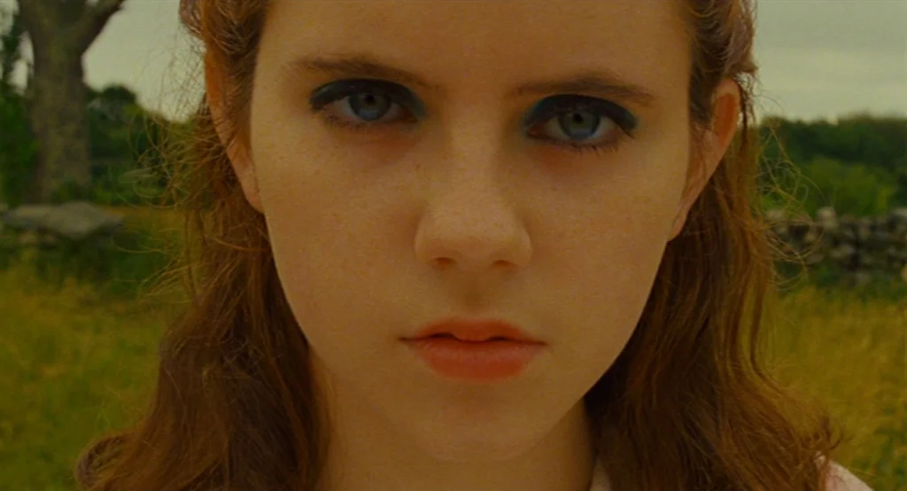 Moonrise Kingdom (2012) movie still: closeup — An open grassy field with a distant tree and stone wall boundary visible. A young girl with blue…