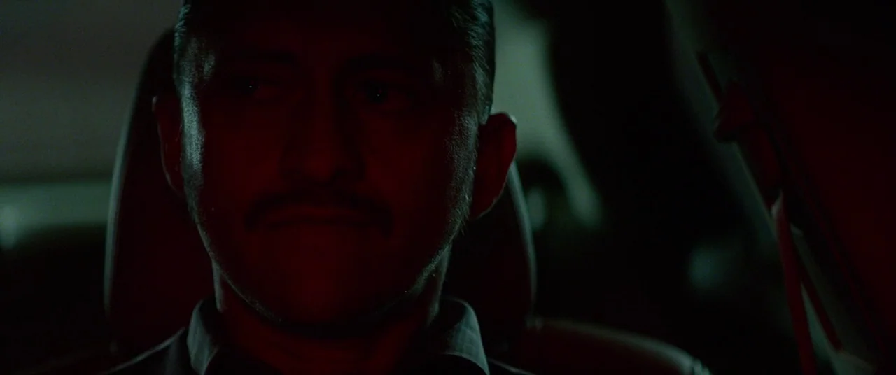 Triple 9 (2016) movie still: closeup — A dark vehicle interior with the silhouette of a headrest visible behind the subject. A man sits behind the…