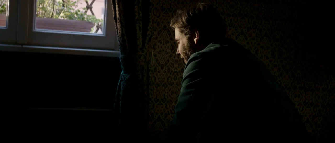 The Face of an Angel (2014) movie still: medium — A dimly lit room with ornate, dark wallpaper features a single window with drawn curtains. A man…