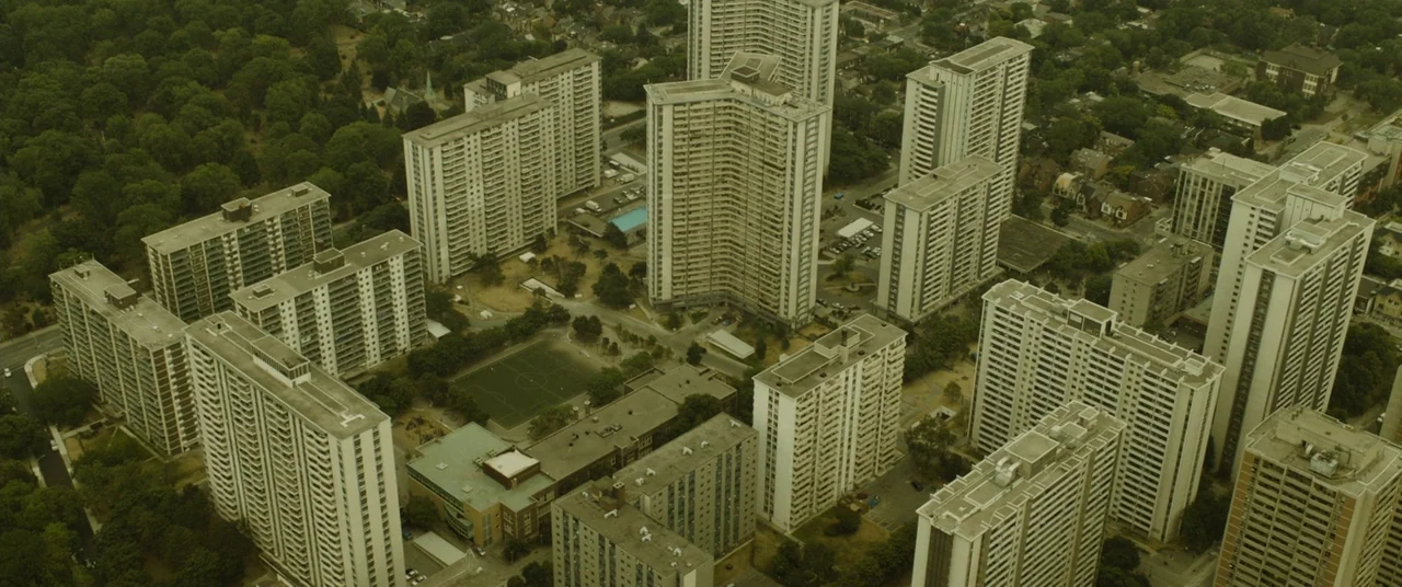 Enemy (2013) movie still: establishing — An aerial view captures a sprawling high-rise residential complex surrounded by dense green trees. Numerous…