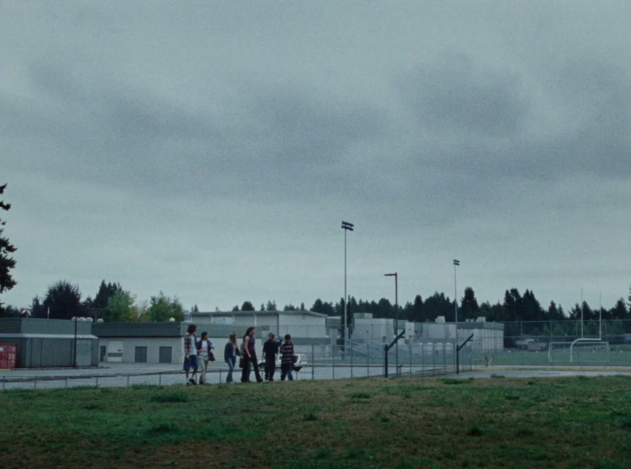 Riceboy Sleeps (2022) movie still: wide — A group of teenagers walks along a school perimeter under an overcast gray sky. Tall stadium lights and…