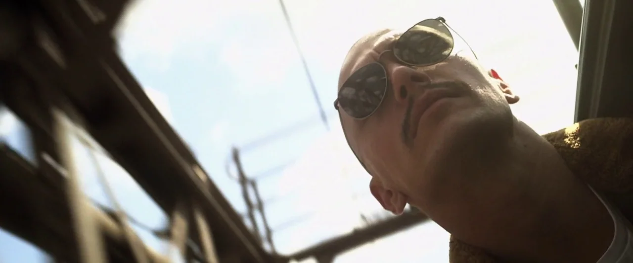 Zeroville (2019) movie still: closeup — An open window frame reveals a glimpse of power lines against the blue sky. A man with a shaved head and…