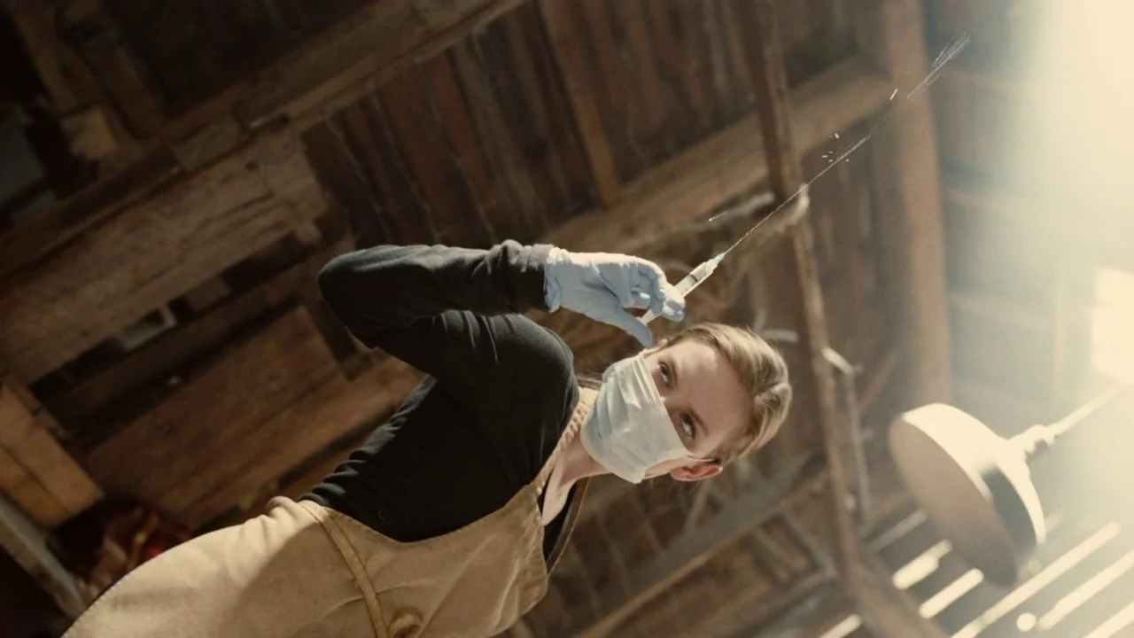 Splice (2009) movie still: medium — A woman stands in a wooden-beamed workshop with rough textures and rustic fixtures. She holds a syringe to the…