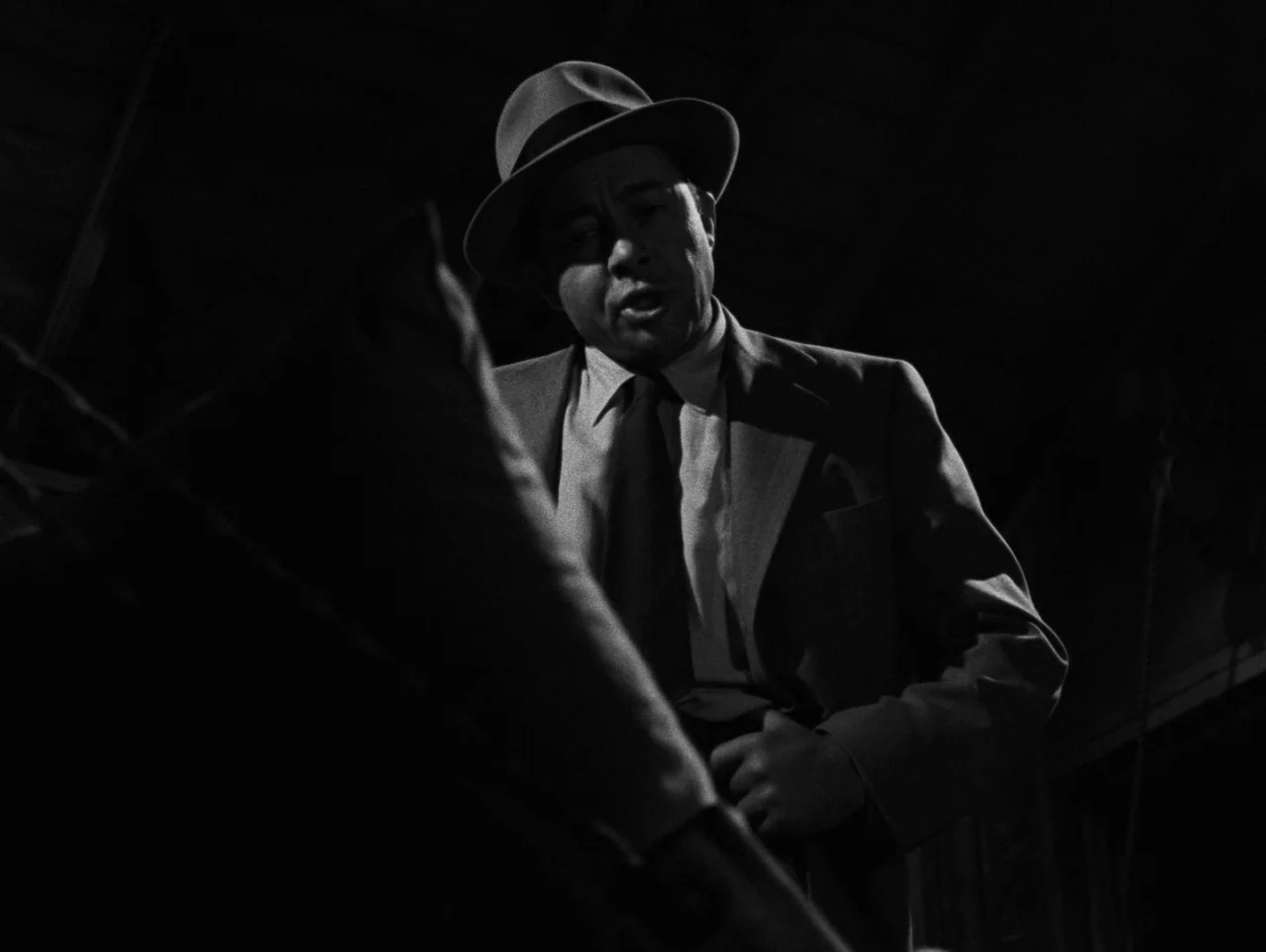 Pickup on South Street (1953) movie still: medium — A dark attic or enclosed space features a man standing in deep shadows. The man wearing a fedora…