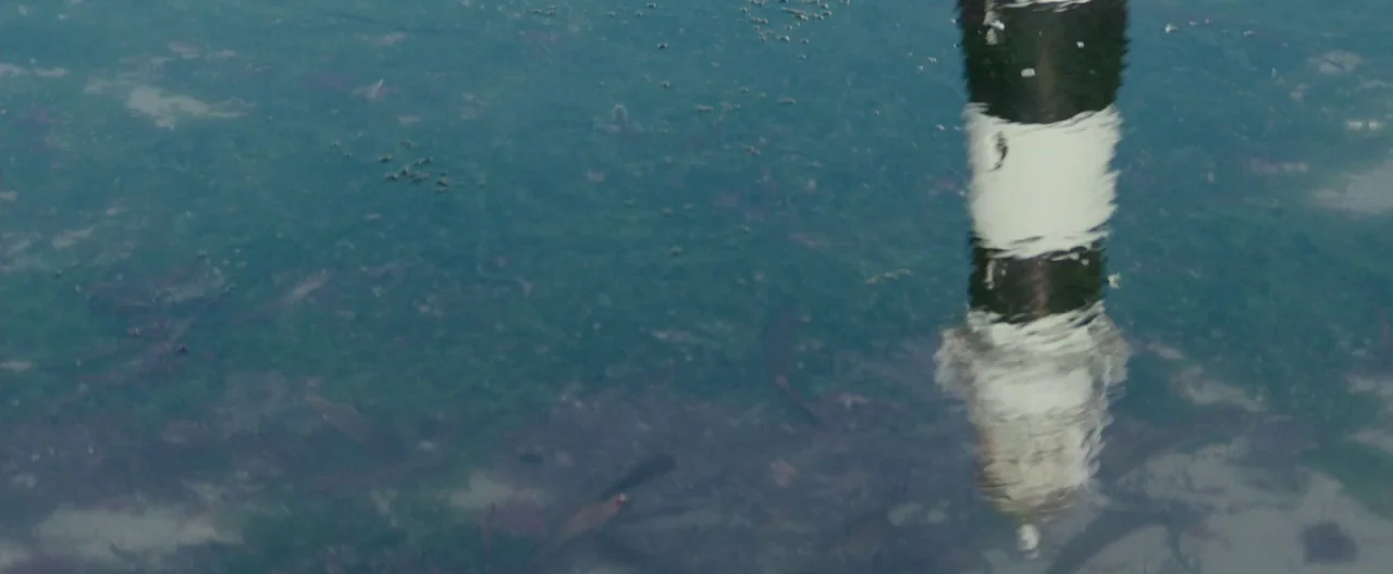 The Light Between Oceans (2016) movie still: establishing — A body of water shows a distorted reflection of a black and white structure. The water…