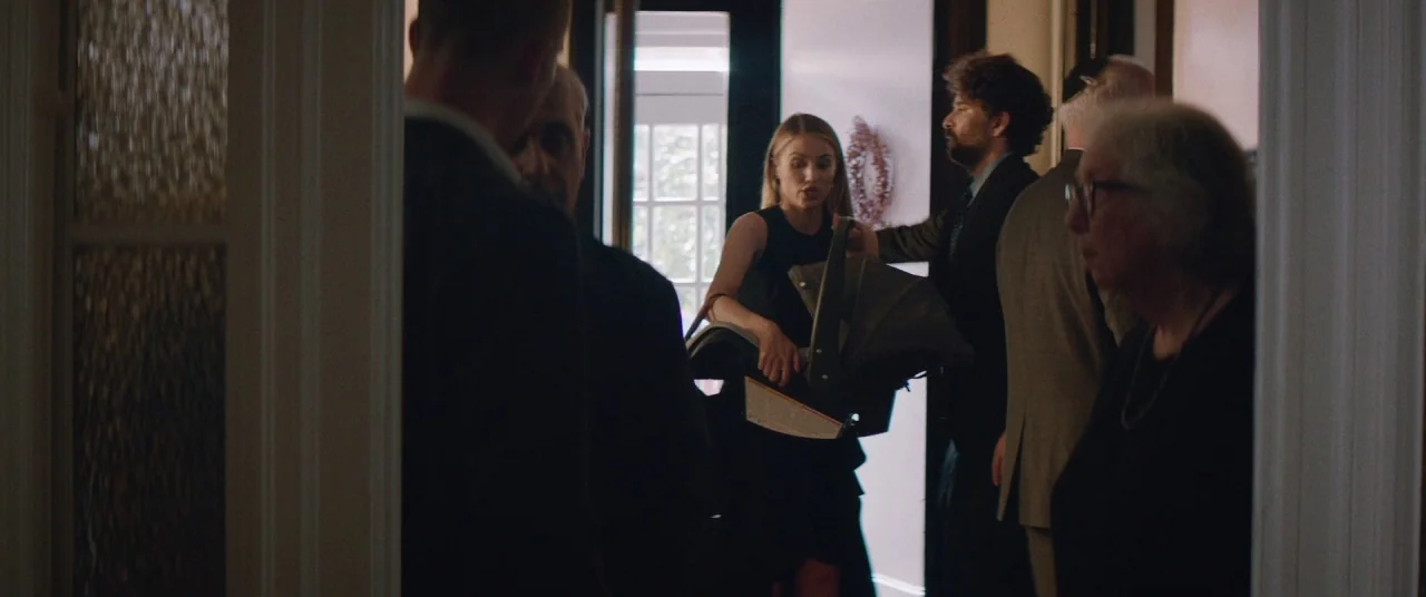 Shiva Baby (2020) movie still: medium — A group of people in formal attire gather in a dimly lit residential hallway. A woman walks toward the…