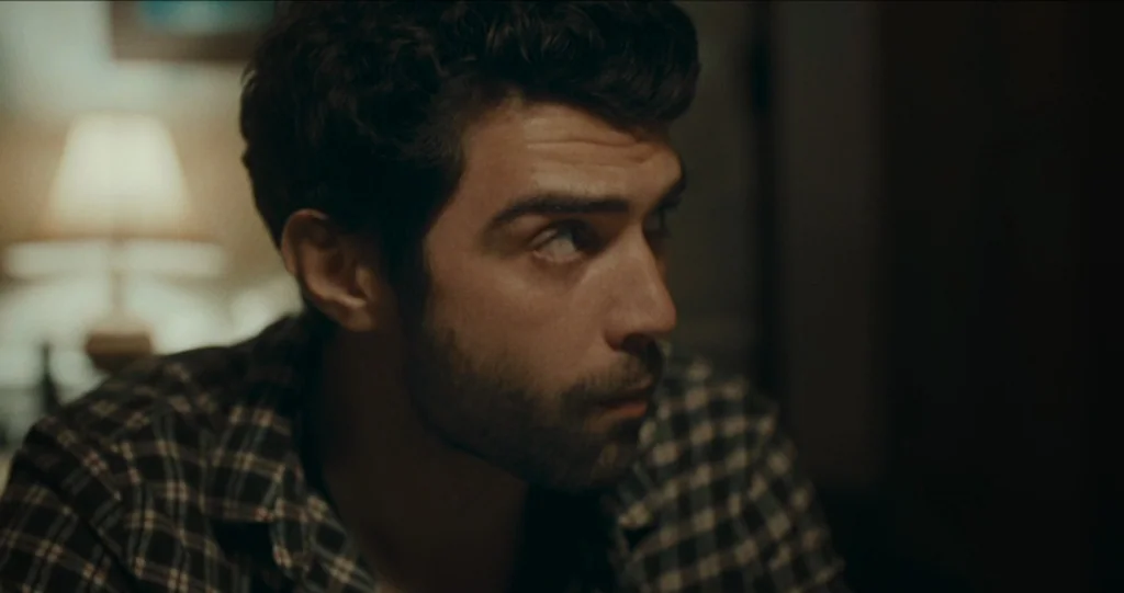 God’s Own Country (2017) movie still: closeup — An indoor space with dim lighting and a blurred lamp in the background. A man with curly dark hair…