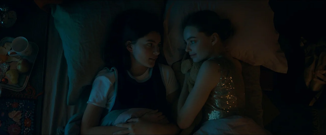 Birds of Paradise (2021) movie still: medium — Two young women lie on a bed at night within a dark bedroom. They face each other while resting on…