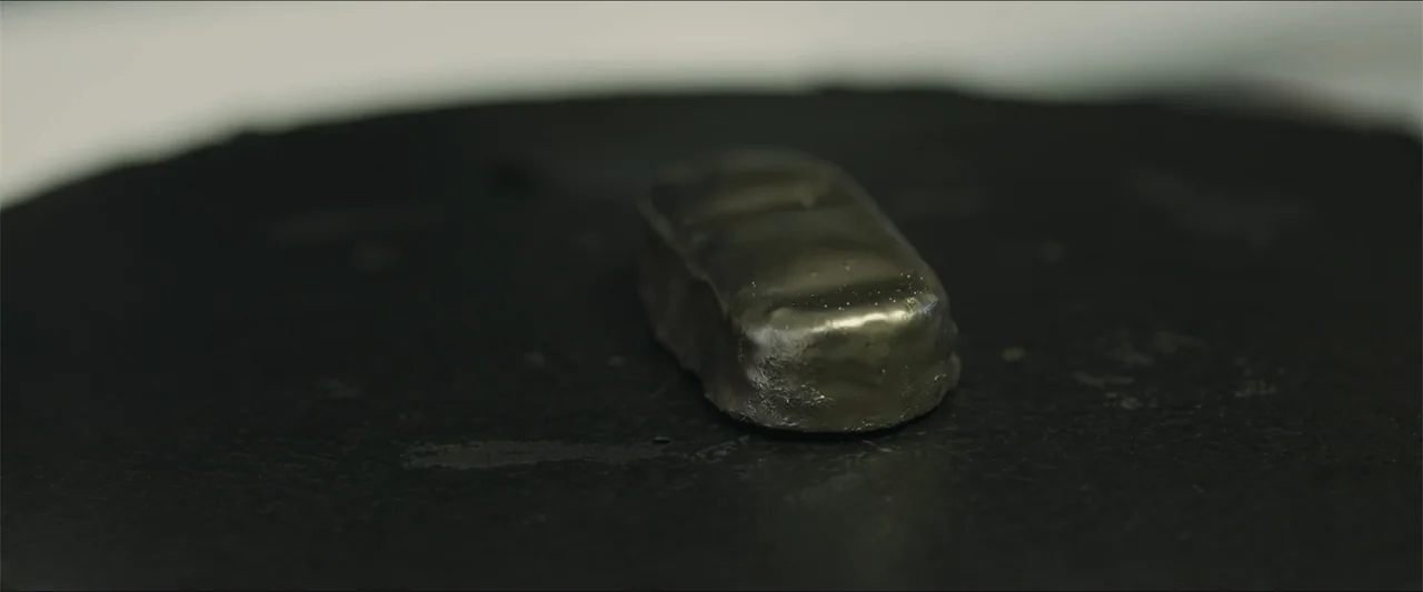 Heavenly Deposit (2019) movie still: closeup — A small, rectangular, metallic-looking object rests on a circular, dark, textured stone surface. The…