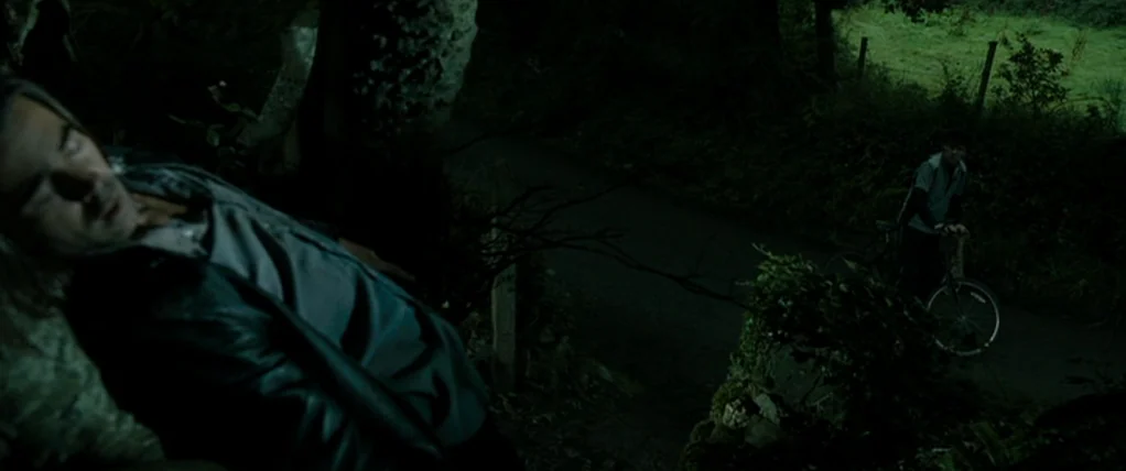Ondine (2009) movie still: medium — A dimly lit country road bordered by overgrown vegetation and a wire fence. One man reclines on the ground while…