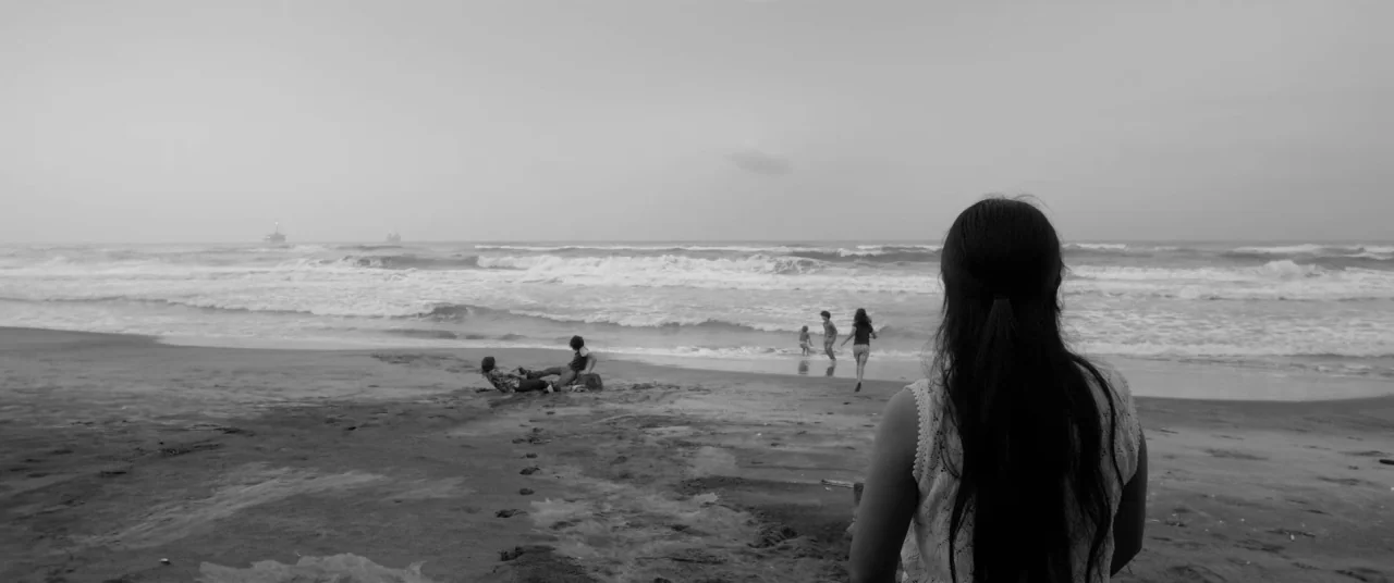 Roma (2018) movie still: wide — A wide beach under a grey overcast sky with waves crashing against the sand. A woman stands in the foreground…