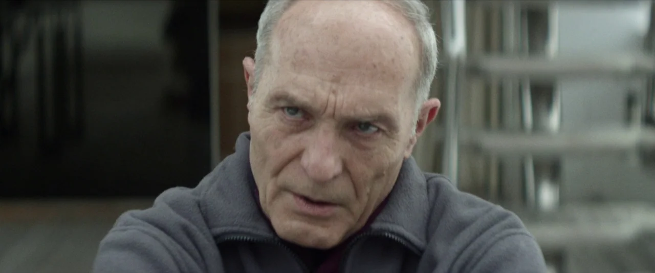 Chevalier (2015) movie still: closeup — An interior space provides a blurred background with metallic or structural elements visible. An older man…