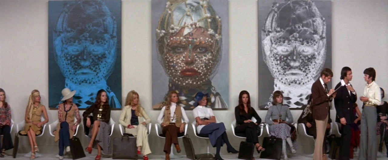 Klute (1971) movie still: wide — A long white wall features three large paintings of faces adorned with intricate beadwork patterns. A group of…