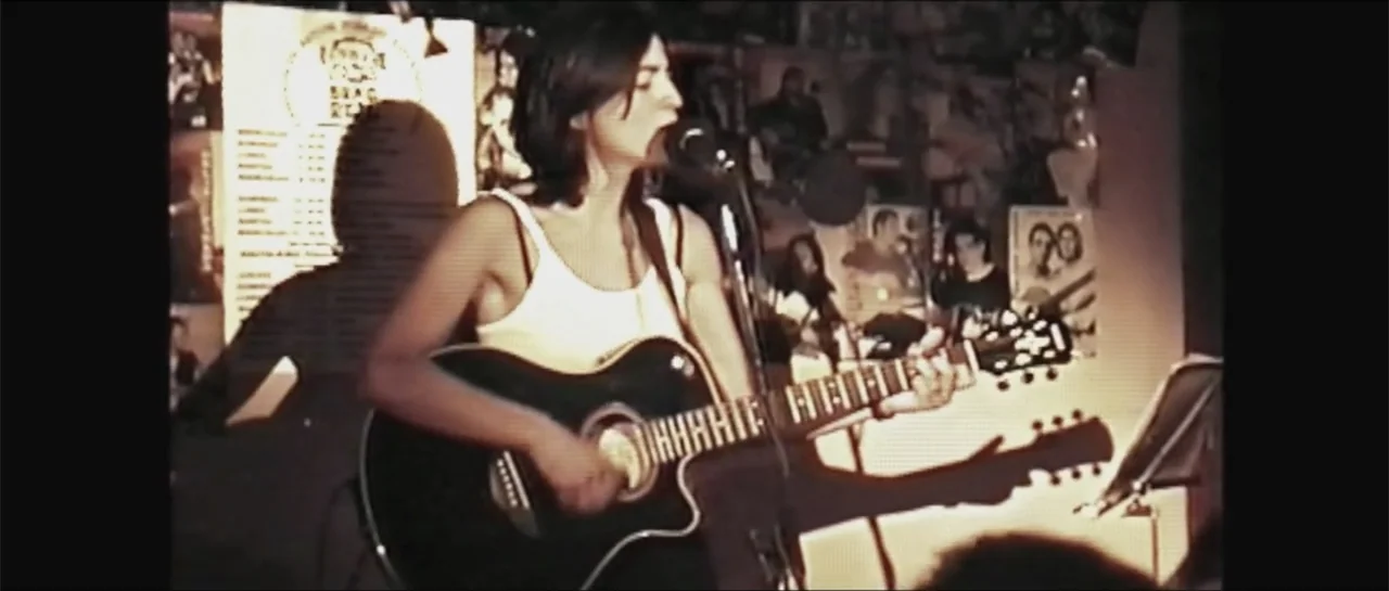 10 Years with Bebe (2016) movie still: medium — A dimly lit live music venue features walls covered in posters and flyers. A female musician stands…