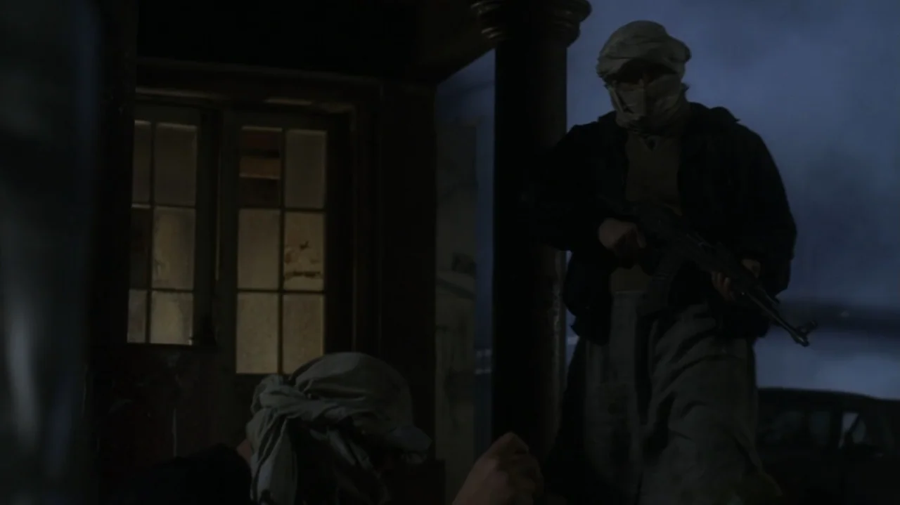 The Shepherd (2023) movie still: medium — Two individuals stand on a dark, shadowed porch at night near a paneled door. One man holds a rifle while…