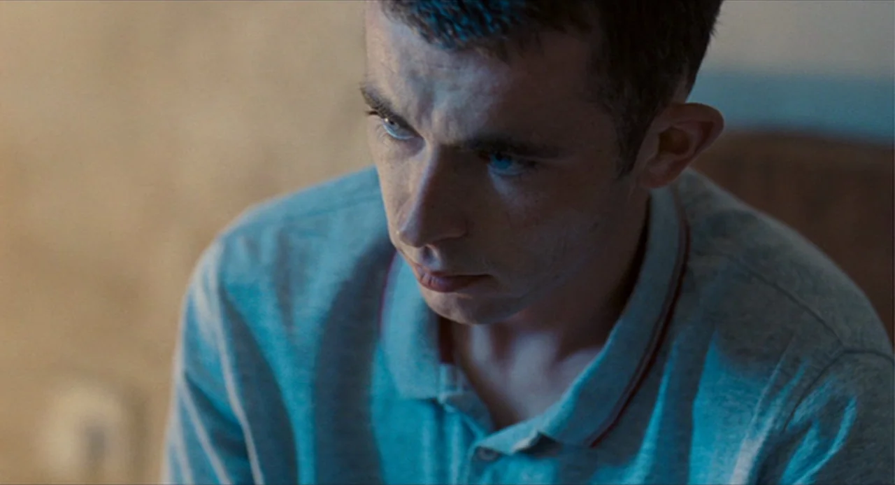 The Angel’s Share (2012) movie still: closeup — An indoor space features a wall with textured wallpaper behind a young man. The man looks downward…