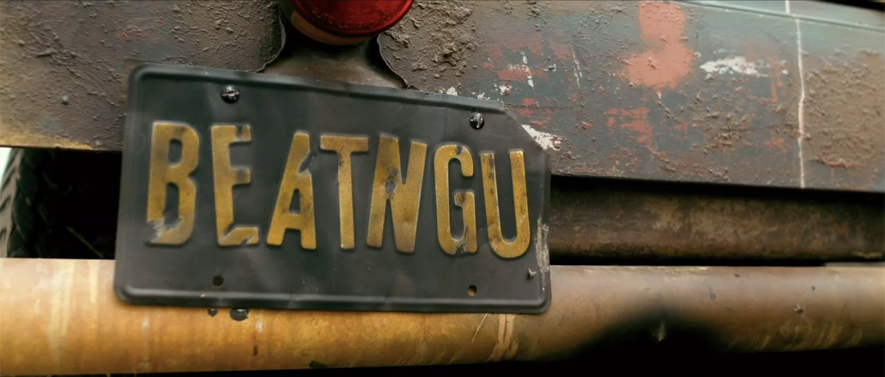 Jeepers Creepers 3 (2017) movie still: closeup — A close-up view of a mud-caked metal car bumper featuring a weathered license plate. The black…