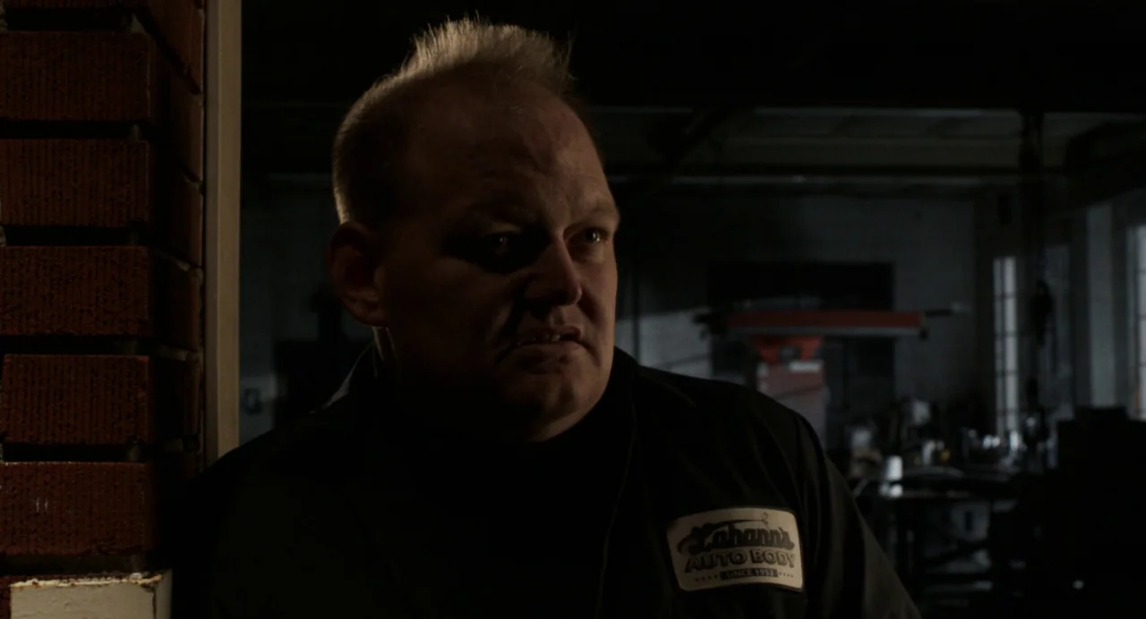 Happer’s Comet (2022) movie still: medium — A dimly lit industrial auto shop interior with dark walls and red brick textures. A man wearing a work…