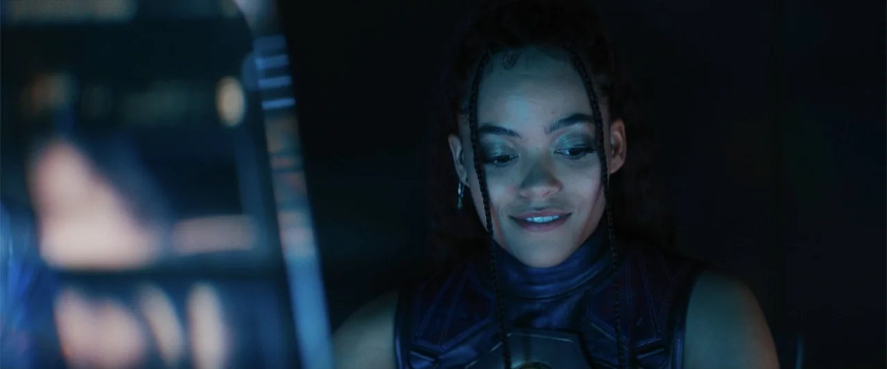 Black Adam (2022) movie still: closeup — A dark, dimly lit cockpit interior is dominated by a glowing digital control console. A young woman with…