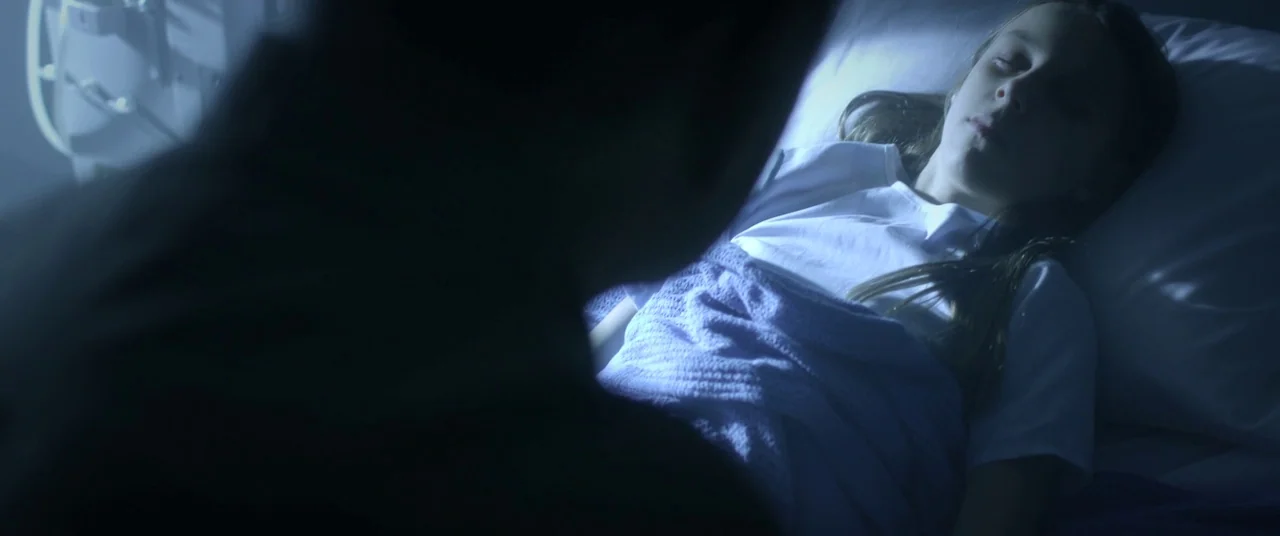 The Void (2016) movie still: medium — A dark, blue-toned room features a girl resting on white pillows under a blanket. A silhouetted figure stands…