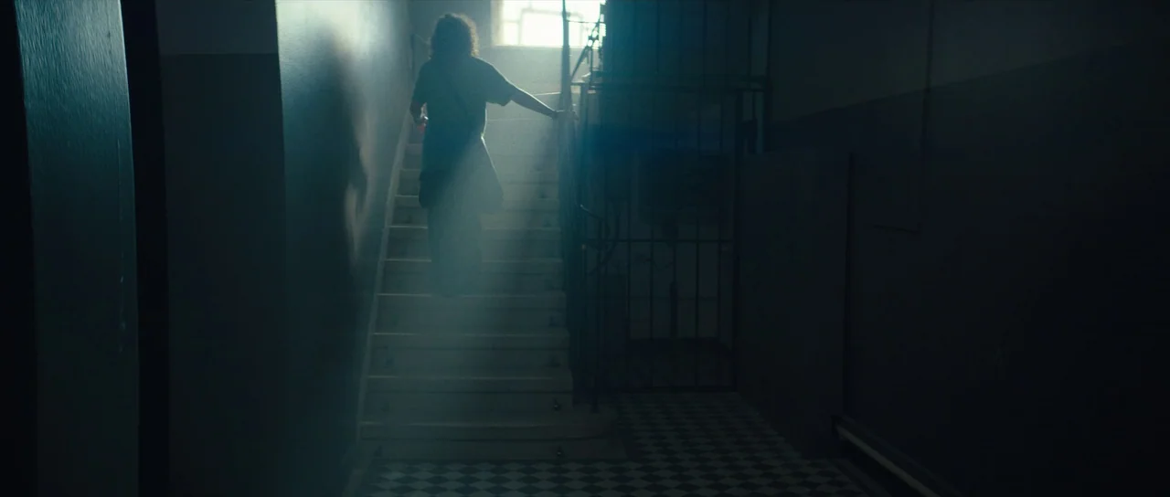 Hellraiser (2022) movie still: fullbody — A dimly lit industrial stairwell features a checkered tiled floor and metal railings. A person in dark…