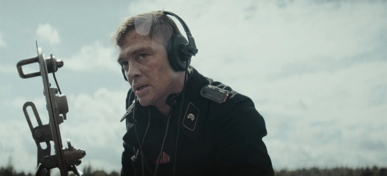 The Tank (2025) movie still: medium — A man in a black 1940s military uniform operates a metallic device outdoors. He wears heavy black headphones…