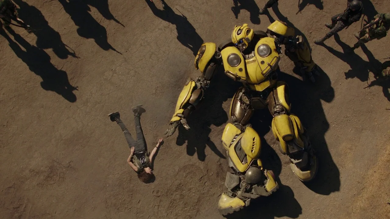 Bumblebee (2018) movie still: wide — A vast, dry desert floor covered in light brown dirt and small pebbles. A giant yellow robotic figure stands…