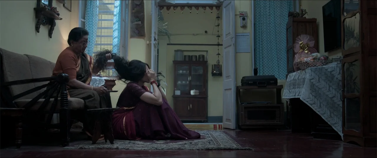 Cheekatilo (2026) movie still: medium — A domestic interior with a vintage wooden cabinet, a beige sofa, and patterned flooring. An older woman sits…