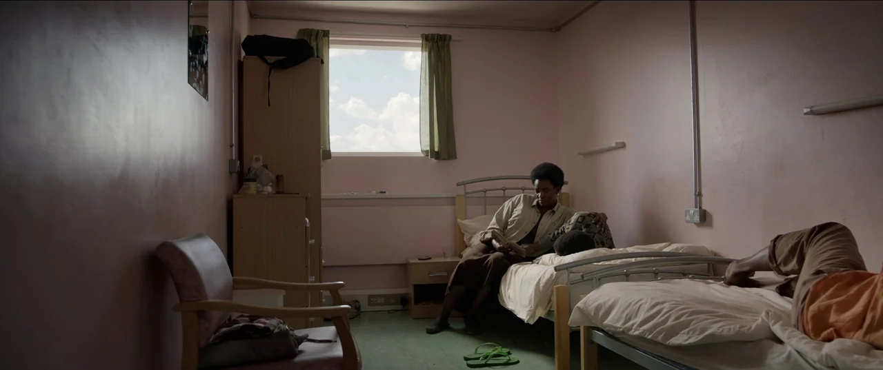 His House (2020) movie still: wide — A spare hospital room with pink walls and twin metal-framed beds. A woman reads a book while sitting beside a…