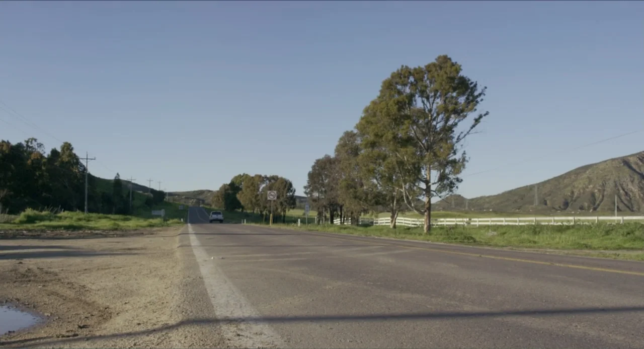 Instantes Capturados (2022) movie still: establishing — A quiet two-lane highway stretches across a rural landscape with sparse trees and hills. A…
