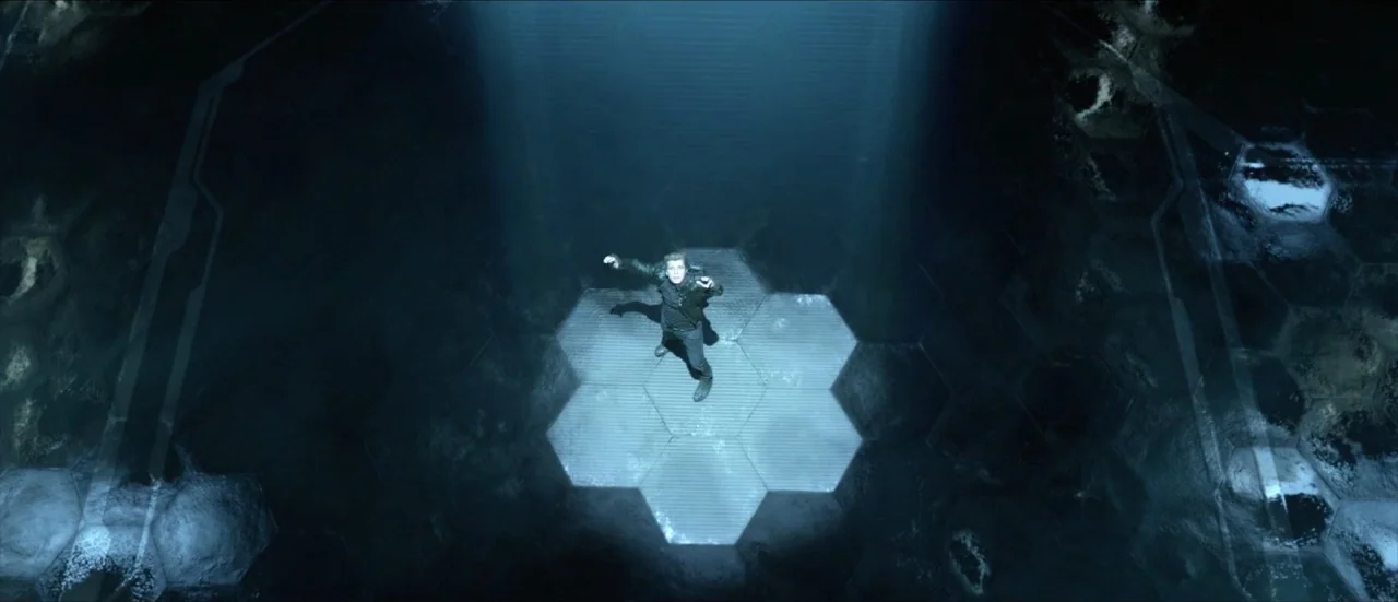 TRON: Legacy (2010) movie still: wide — A dark, hexagonal grid floor stretches across a vast, industrial, futuristic space. A lone young man stands…