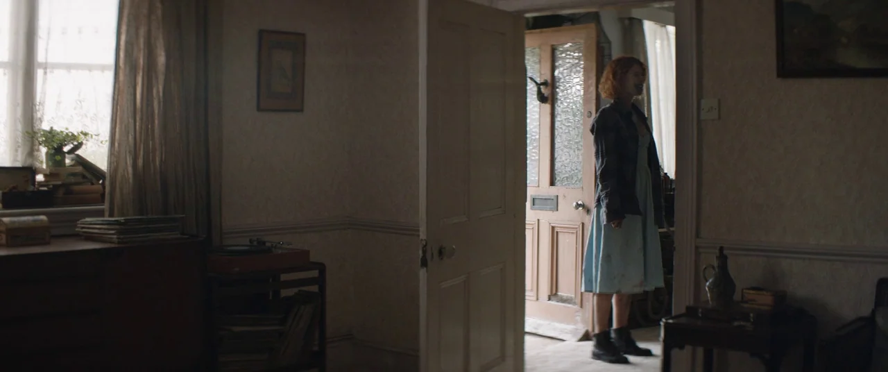 Beast (2017) movie still: fullbody — A residential hallway with patterned wallpaper and an open door leading outside. A young woman with short…