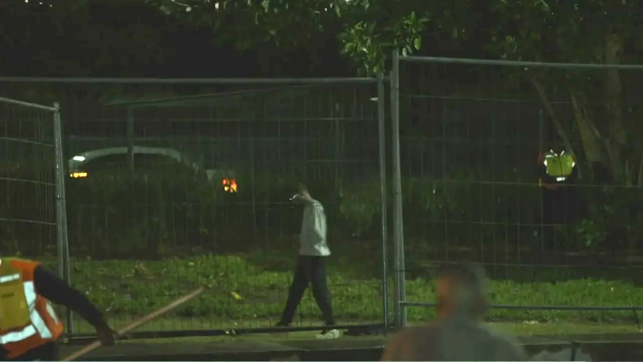 African Throne World Tour (2024) movie still: wide — A man walks past a wire construction fence at night in an urban area. He wears a light-colored…