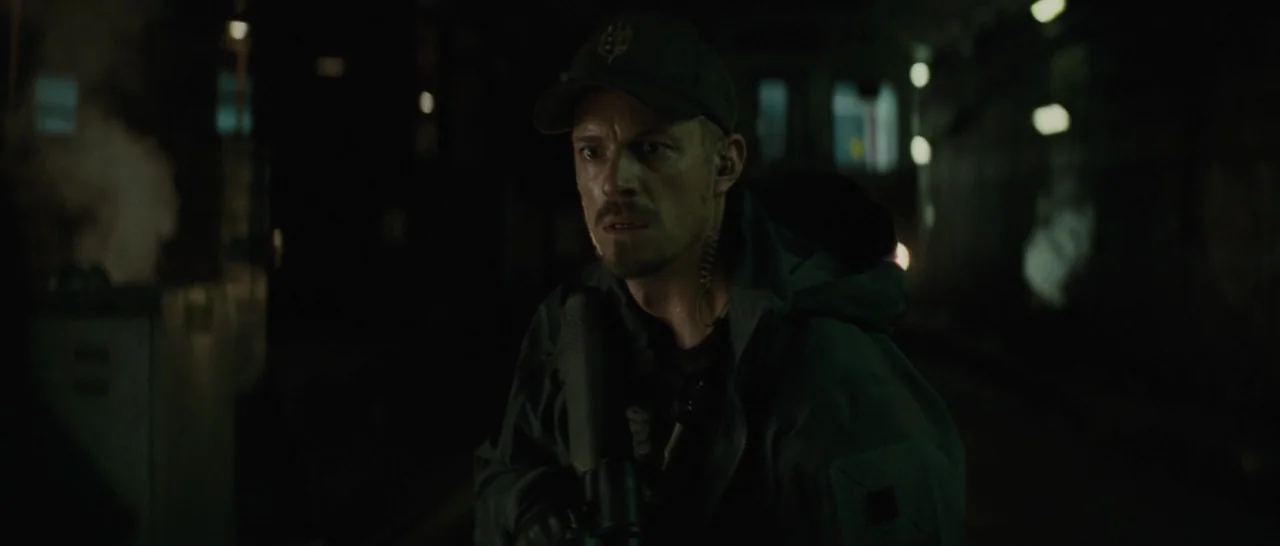 Suicide Squad (2016) movie still: medium — A dark, dimly lit subway tunnel with metallic structural elements and overhead lighting. A man in…