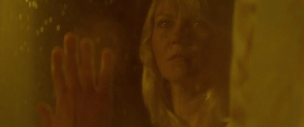 Woodshock (2017) movie still: closeup — A person looks through a pane of glass covered in condensation at night. A yellow-toned light source…