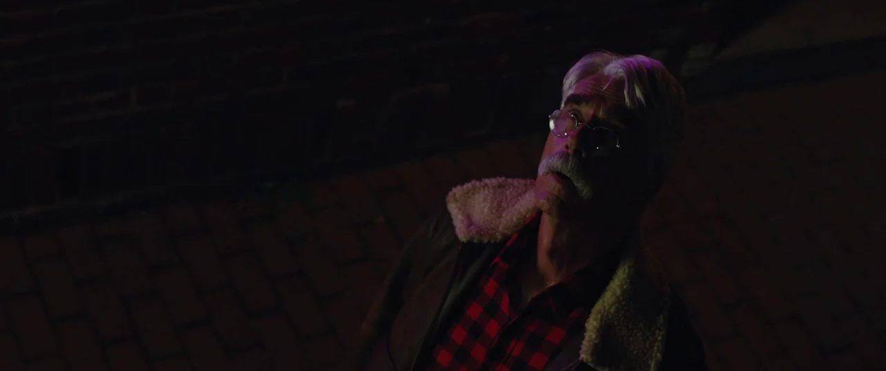 The Man Who Killed Hitler and Then the Bigfoot (2018) movie still: medium — A man stands outdoors at night near a dark red brick wall. The person…