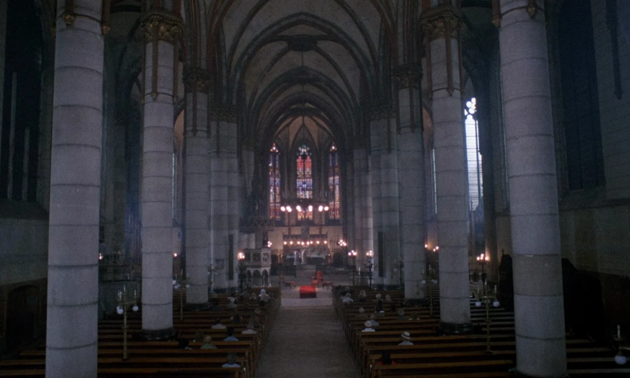 The Church (1989) movie still: establishing — A spacious gothic cathedral nave features towering stone pillars and rows of wooden pews. Dimly lit…