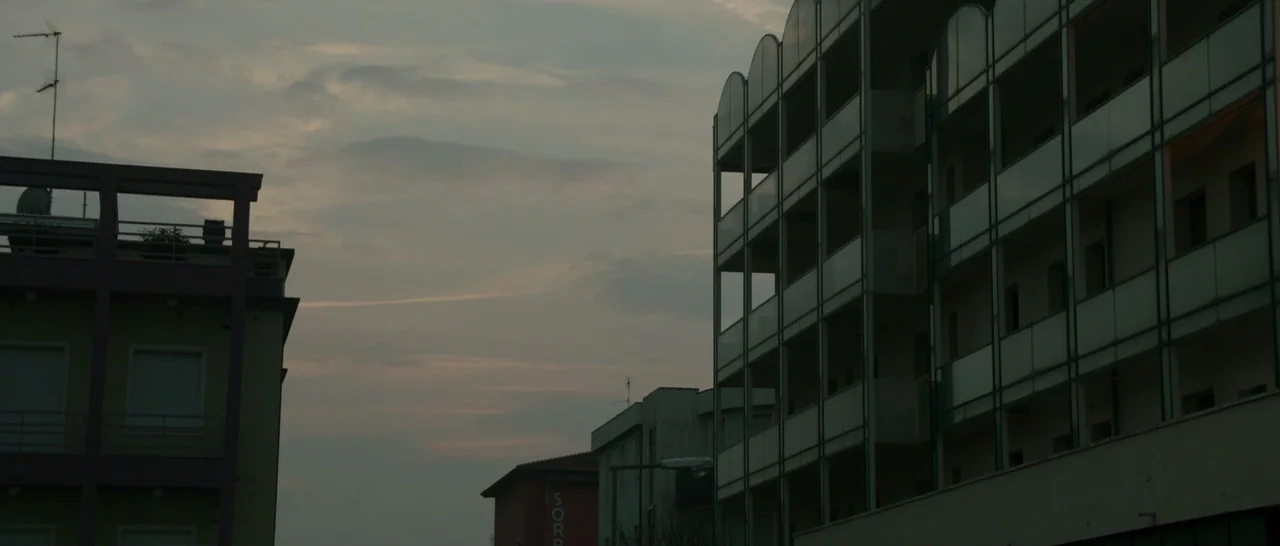 The Face of an Angel (2014) movie still: establishing — Residential buildings flank a narrow gap revealing a dim, overcast twilight sky. A…