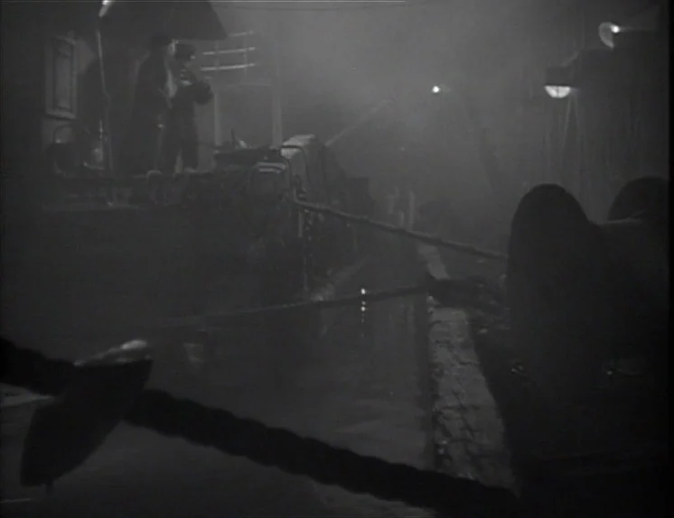 Anna Christie (1930) movie still: wide — A dark, fog-filled maritime area at night with mooring ropes extending into the distance. Two indistinct…