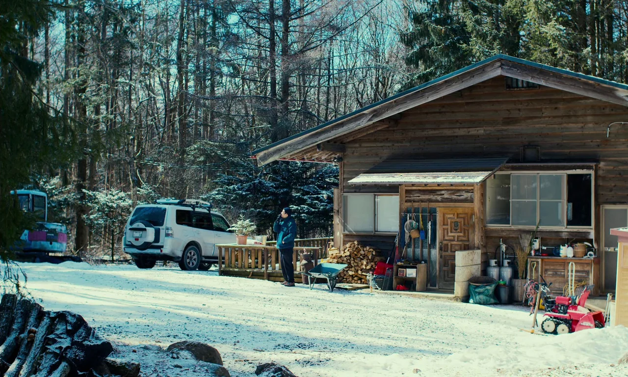 Evil Does Not Exist (2023) movie still: wide — A wooden cabin sits at the edge of a snow-covered clearing near dense woods. A person stands on the…