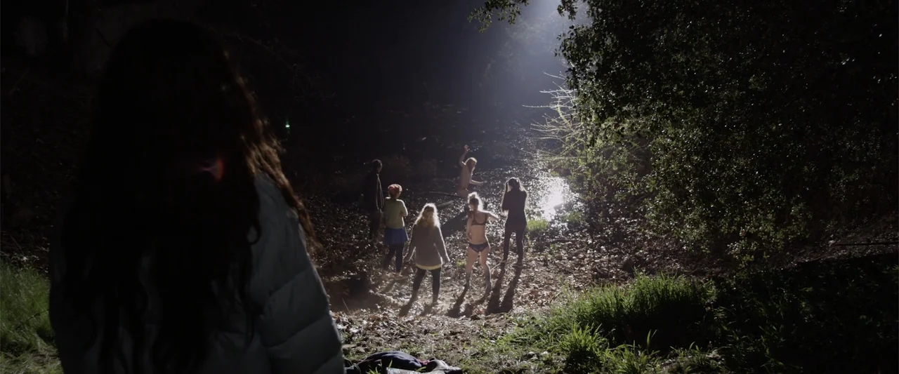 Mermaid Down (2019) movie still: wide — A group of people stands on a leaf-covered bank beside a reflective stream. The view is framed over the…
