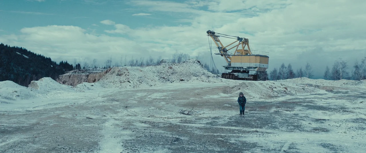 R.M.N. (2022) movie still: wide — A vast, snowy industrial quarry features mounds of gravel and a large yellow crane. A small child walks across the…