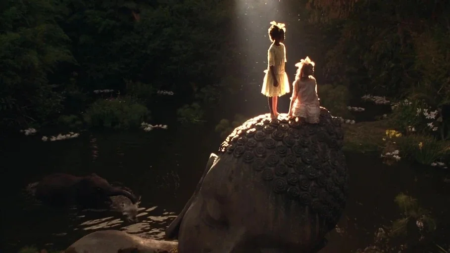 A Little Princess (1995) movie still: wide — Two children stand atop a massive stone Buddha head submerged in a dark pond. An elephant is partially…