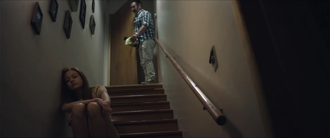 Captive (2020) movie still: medium — A dark residential staircase with a wooden handrail and framed pictures on the wall. A young woman sits on the…