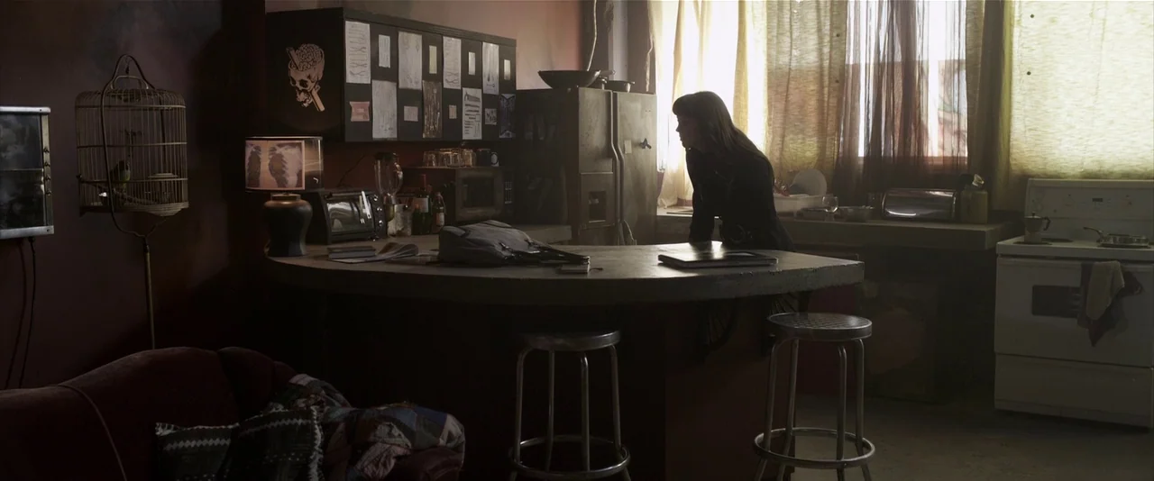 American Mary (2012) movie still: wide — A dimly lit room features a central wooden counter, metal stools, and a birdcage. A woman with dark hair…
