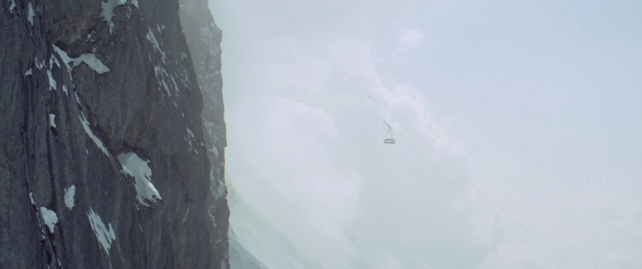 Force Majeure (2014) movie still: establishing — A vertical, jagged rock face covered in patches of snow dominates the left side. A distant cable…